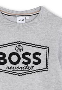 Pull gris à col rond avec un logo noir portant la mention "BOSS seventy". Le matériau semble doux, avec une texture tricotée.