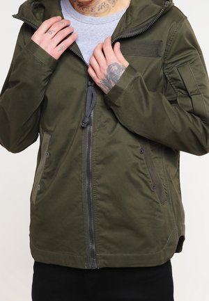 Veste vert olive avec fermeture éclair à l'avant, poignets élastiques et capuche. Dispose de deux poches avant et d'accents texturés sur la poitrine et les manches.