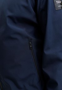 Υφασμα για μπουφάν σε ναυτικό μπλε χρώμα με μαύρη κάθετη τσέπη με φερμουάρ και μια μικρή μαύρη ετικέτα που δείχνει λευκό κείμενο και συντεταγμένες.