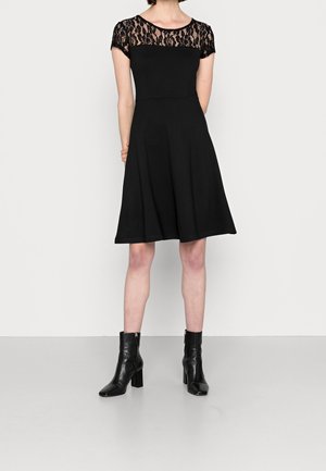 Robe noire avec un corsage ajusté et une jupe évasée. La superposition en dentelle couvre les épaules, ajoutant de la texture. Portée avec des bottines noires.
