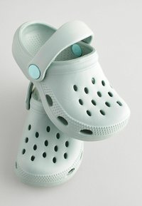 Zoccoli verde menta chiaro con superficie testurizzata, fori di ventilazione, tracolla regolabile e punta arrotondata. Realizzati in materiale flessibile e resistente.