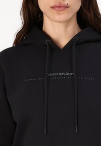 Felpa nera con cappuccio e cordini. Presenta un logo frontale con la scritta "Calvin Klein Jeans" e "Established nineteen seventy-eight" in grigio.