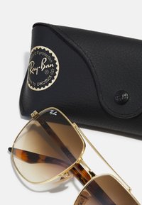 Ochelari de soare Ray-Ban cu ramă metalică aurie și lentile maronii cu gradient, asortați cu o carcasă din piele neagră cu un logo auriu și închidere tip clapă.