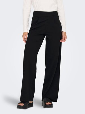 Mujer con pantalones negros de pierna ancha y cintura alta, top blanco de manga larga y sandalias negras de plataforma con punta abierta, de pie sobre un fondo liso y claro.