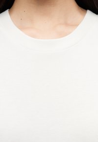 Gros plan sur le cou et le haut de la poitrine d'une personne portant un t-shirt blanc uni à col rond, avec quelques petits grains de beauté sur la peau.