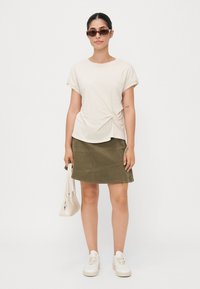 Beige geknoopte t-shirt, olijfgroene corduroy rok, witte sneakers en een lichtbeige handtas; model draagt donkere zonnebril.