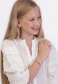 Blouse blanche avec des volants texturés, boucles d'oreilles en argent et bracelet. Le bracelet présente un design épuré et un accent en forme de cœur.