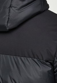 Gros plan de l'arrière d'une veste matelassée noire avec une capuche, montrant différentes textures de tissu et des détails de couture.