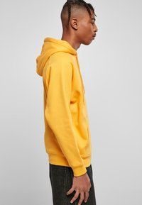 Sudadera amarilla con cordón, bolsillo tipo canguro y puños acanalados. Estilizada con jeans oscuros, vista de lado sobre un fondo claro.
