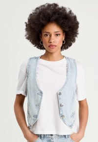Gilet en denim rayé bleu clair avec un col rond, quatre boutons à l'avant et des poches latérales, porté sur un T-shirt blanc.