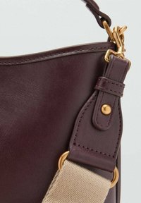 Sac à main en cuir bordeaux avec une texture lisse, présentant un accessoire en métal doré et une bandoulière en toile beige.