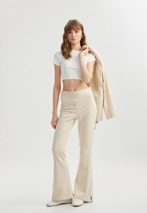 Afskåret hvid t-shirt, beige klokkeformede bukser med høj talje, og en beige blazer holdt over skulderen. Model står mod en lys baggrund.