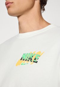 Hvid bomulds t-shirt med en grøn og gul grafik "NIKE" logo med en tekstureret finish. Rund halsudskæring og korte ærmer.