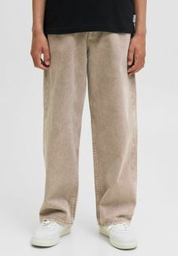 Bež hlače širokih nogavic iz mehke, teksturirane tkanine. Imajo vidno šivanje in visok pas, kombinirane z belimi športnimi copati.