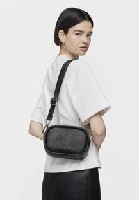 Czarna skórzana torba na ramię z teksturowanym wykończeniem, złotymi akcentami i wytłoczonym logo z przodu. W zestawie regulowany pasek.