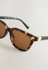 Bruine schildpadprint zonnebril met een geometrisch montuur, bruine glazen en gouden accenten op de pootjes. Gladde textuur en modern design.