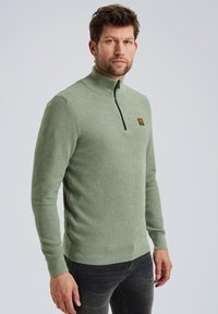 Groene ritsvest met een gestructureerde stof en lange mouwen; heeft een zwarte rits en een klein logo-opdruk op de borst.