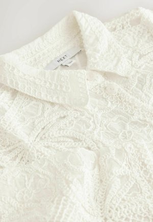 Hvid bluse med et tekstureret design, der har en krave og to knapper. Indviklede blondeagtige mønstre over hele stoffet.