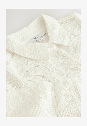 Hvid bluse med et tekstureret design, der har en krave og to knapper. Indviklede blondeagtige mønstre over hele stoffet.