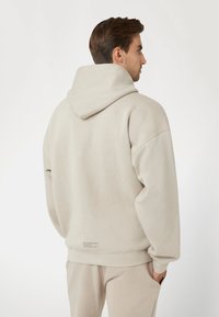 Šviesiai ruda gobtuvinė džemperis su laisva siluetu, turintis akcentuojamą gobtuvą, kengūros tipo kišenę ir raukšlėtus rankogalius. Etiketė matoma šalia apačios.