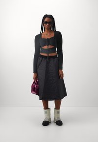 Crni rebrasti crop top s detaljem vezanja sprijeda, u kombinaciji s crnom šivnom suknjom, bijelim čizmama i sjajnom ružičastom torbom. Uključene sunčane naočale.