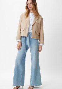 Blazer beige cropped con due tasche frontali, abbinato a jeans a gamba larga blu chiaro con cintura in tessuto. Il modello indossa tacchi.