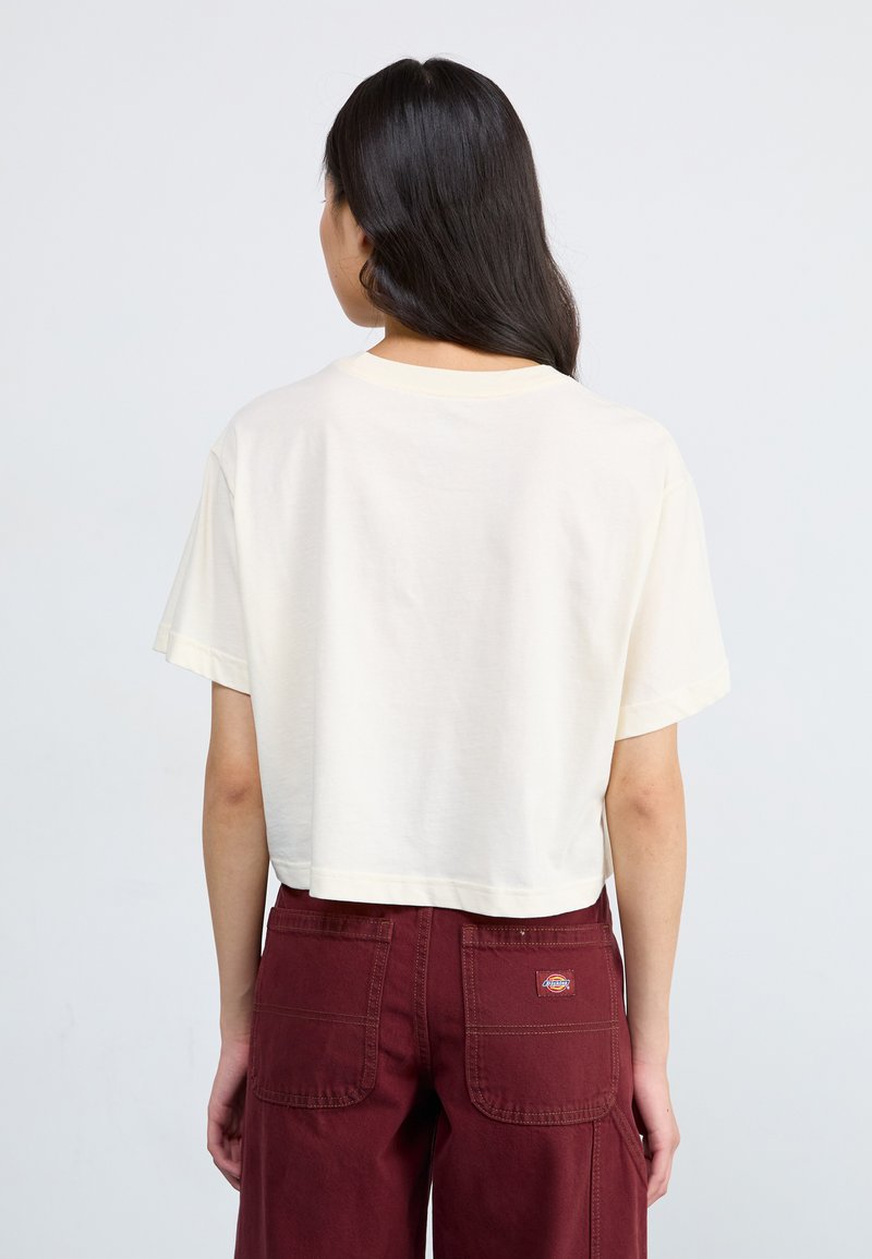Camiseta corta de manga corta en color crema. Tejido suave con un diseño sencillo. Combinada con pantalones burdeos oscuros que tienen dos bolsillos traseros.