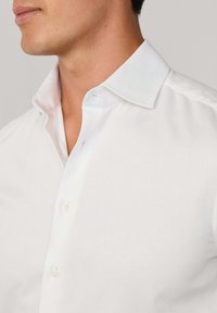 Uomo che indossa una camicia bianca con colletto aperto, due bottoni abbottonati, mostrando una texture di tessuto pulita e liscia.