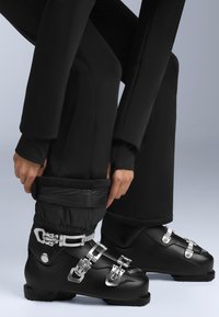 Czarne buty narciarskie z srebrnymi klamrami, o gładkim wykończeniu i zaokrąglonym czubku. Osoba dostosowuje czarne spodnie narciarskie z elastycznymi mankietami.
