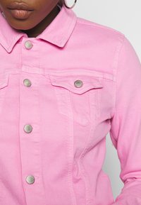 Chaqueta de denim rosa con diseño clásico de cuello, broches de presión plateados y dos bolsillos en el pecho con solapas. Textura suave y costuras detalladas.