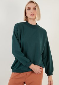 Felpa oversize color teal con collo alto e polsini a coste. Tessuto morbido con vestibilità rilassata, abbinata a pantaloni color marrone chiaro.