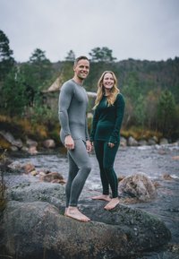 Zwei Personen stehen auf einem Felsen am Fluss und tragen enganliegende thermische Unterwäsche: eine hat ein hellgraues Oberteil und Leggings an, die andere ein dunkelgrünes Oberteil und Leggings.