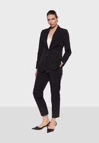 Black suit featuring a fitted blazer with a single button, gold accents, and slim trousers. Paired with pointed-toe black heels.