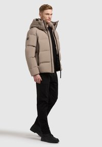 Beige gewatteerde jas met capuchon, zwarte rits en zijzakken. Draagt over een zwart shirt en gecombineerd met zwarte broek en schoenen.
