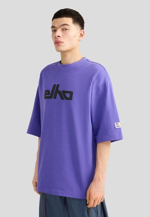 Mladý muž v volném fialovém tričku Elko s černým logem stojí před jednoduchým světle šedým pozadím.