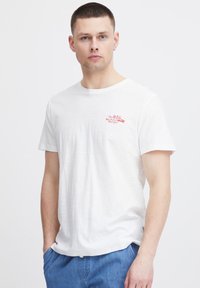 Camiseta de algodón blanca con cuello redondo, que presenta el texto rojo "The BIG Waikiki Grill Burger" en la parte superior izquierda del pecho.