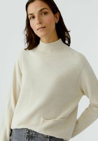 Beiger Pullover mit hohem Kragen aus weichem Material, ausgestattet mit einer Kängurutasche, langen Ärmeln und einem lockeren Schnitt. Minimalistisches Design mit subtiler Textur.