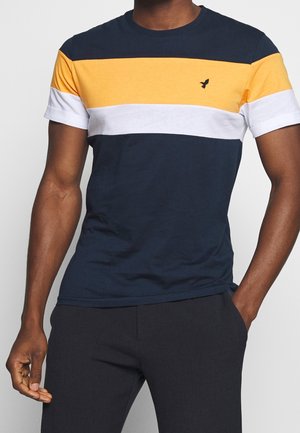 Man draagt een navyblauw, wit en geel t-shirt met korte mouwen en kleurvlakken, met een klein zwart vogel-logo op de borst.