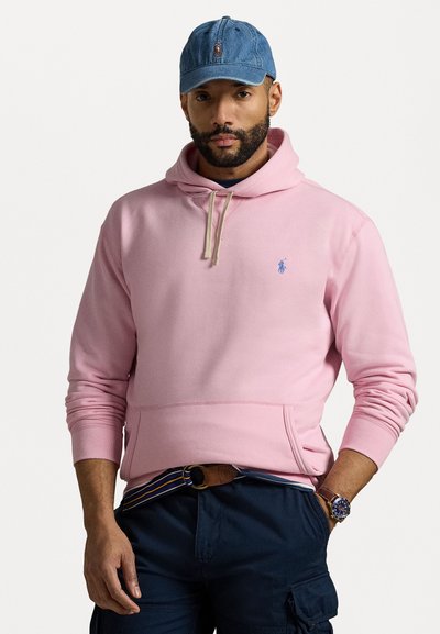 Sweat-shirt à capuche rose avec une poche avant, cordons de tirage beige clair et petit logo bleu. Porté avec un pantalon cargo noir et une casquette en denim.