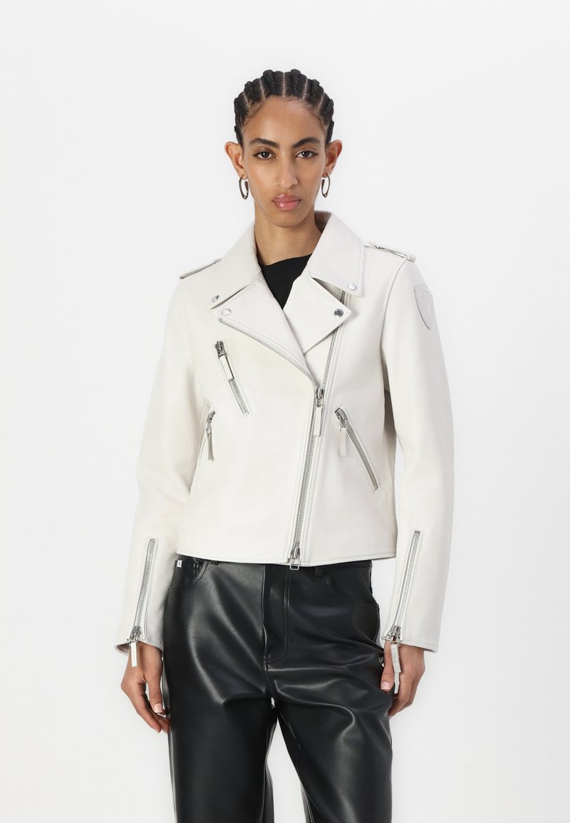 Giacca da moto in pelle bianca con zip argentate, rever inclinati e tasche con zip. Indossata sopra una maglietta nera e abbinata a pantaloni neri.
