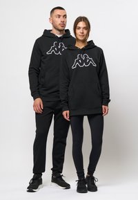 Zwarte hoodies met een kangoeroezak, featuring een wit Kappa-logo. Modellen dragen zwarte joggers en leggings, met zwarte sneakers.