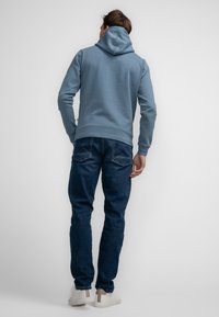 Modrá mikina s kapucí a hladkým povrchem, doplněná tmavě modrými džínami. Bílé tenisky s béžovými detaily. Model zobrazen zezadu.