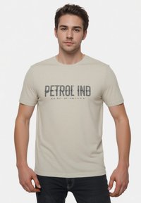 Lys beige T-shirt lavet af blødt stof med rund halsudskæring. Har "PETROL IND" trykt i mørkegrå over brystet. Korte ærmer.