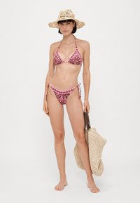 Bikini w kwiatowy wzór w odcieniach różu i brązu, z trójkątnym topem i spodenkami z wiązaniem z boku. W komplecie słomkowy kapelusz i pleciona torba.