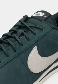 Nike tenisice s tamnozelenim nubuk gornjim dijelom, sivim kožnim swooshom, crnim oznakom na jeziku i tamnim vezicama s teksturiranim detaljima.