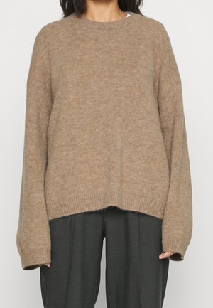 Kobieta ubrana w oversize'owy beżowy sweter z dzianiny z prążkowanymi mankietami i ściągaczem, w połączeniu z luźnymi ciemnoszarymi spodniami.