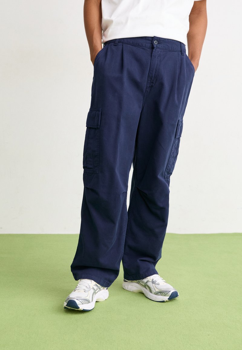 Pantaloni cargo bleumarin, cu o croială lejeră, având multiple buzunare laterale și închidere cu nasture, confecționați dintr-un material neted și ușor.