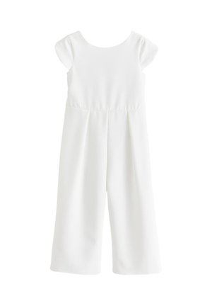 Fiatal lány fehér ruhában, aki egy csokor sárga és fehér virágot tart bézs háttér előtt.