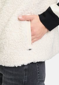 Giacca in pile bianca e testurizzata con una mano in una tasca laterale. Presenta maniche nere con strisce bianche e un design a vestibilità rilassata.