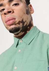 Lys grøn, skjorte med knapper og krage, laget av et teksturert stoff. Har en enkelt brystlomme og to-knapps placket.